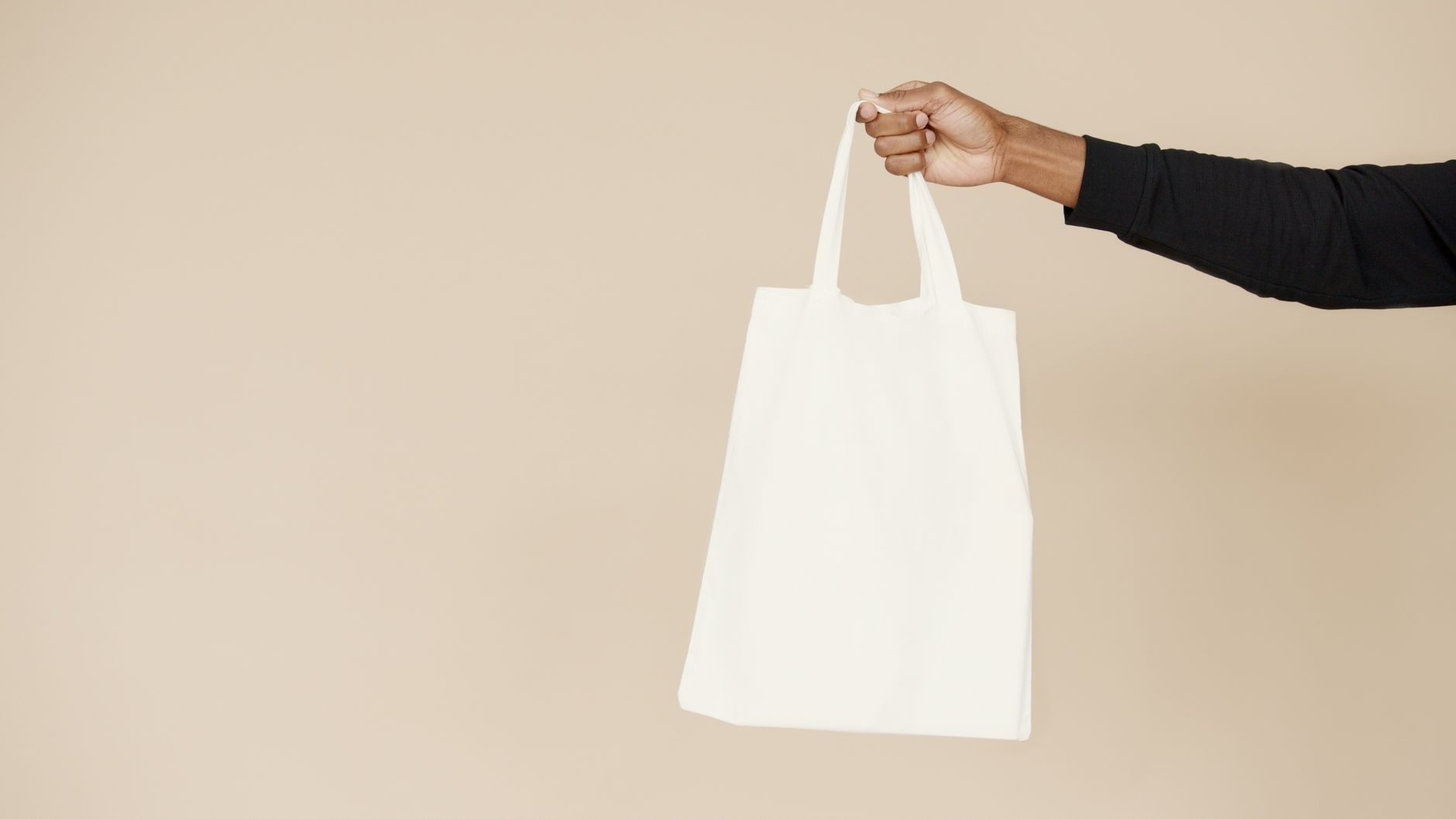 En hand håller upp en tom vit tygkasse mot beige bakgrund